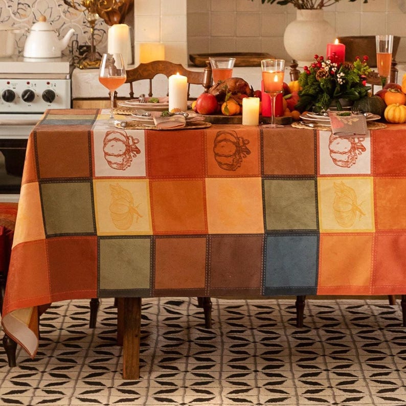 May include: Rectangular tablecloth with warm orange, brown, yellow, and green patchwork blocks, featuring pumpkin and leaf motifs. A festive dining table is set with candles, fruit, drinks, and plates.