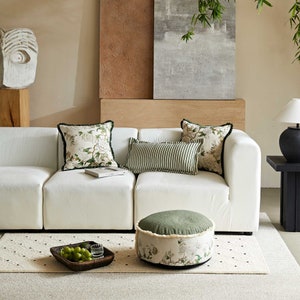 May include: A white sectional sofa with green and white patterned throw pillows and a green and white patterned ottoman. The sofa is in a living room with a beige wall and a painting on the wall behind it.