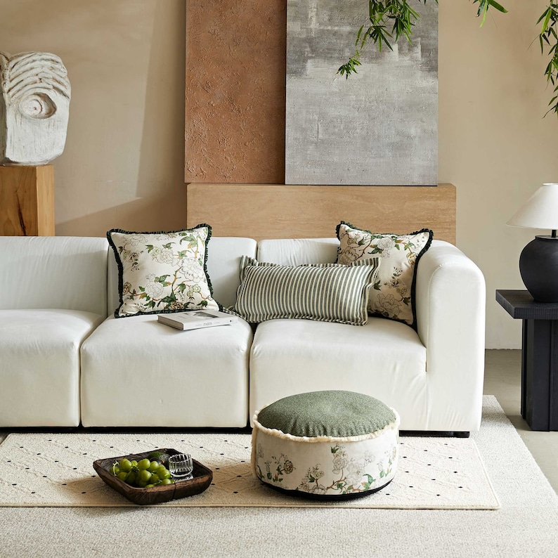 May include: A white sectional sofa with green and white patterned throw pillows and a green and white patterned ottoman. The sofa is in a living room with a beige wall and a painting on the wall behind it.