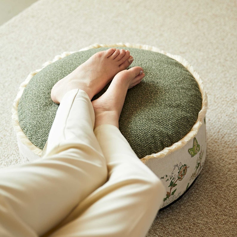 May include: A round green and white ottoman with a floral pattern and a white fringe. A person is sitting on the ottoman with their legs crossed.