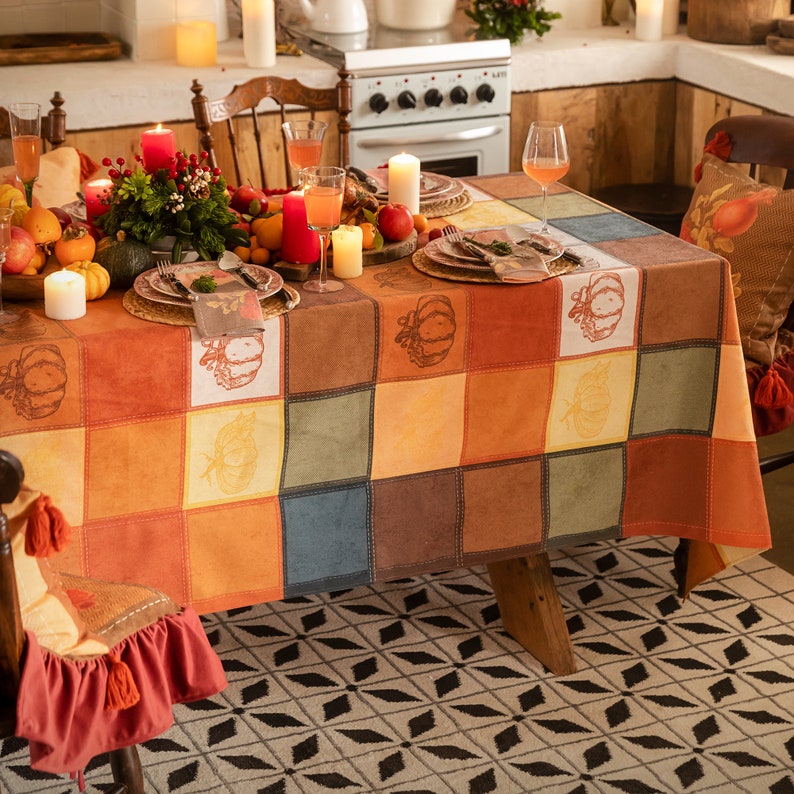 May include: A festive table setting with a patchwork tablecloth featuring pumpkin designs. The table is set with plates, napkins, and glasses, and is decorated with candles, fruit, and flowers.