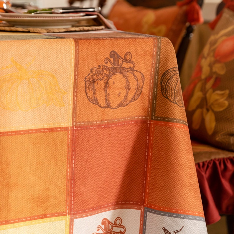 May include: Close-up of a fall tablecloth with orange patchwork blocks and pumpkin illustrations. The fabric drapes over the table edge with warm autumn decor in the background.