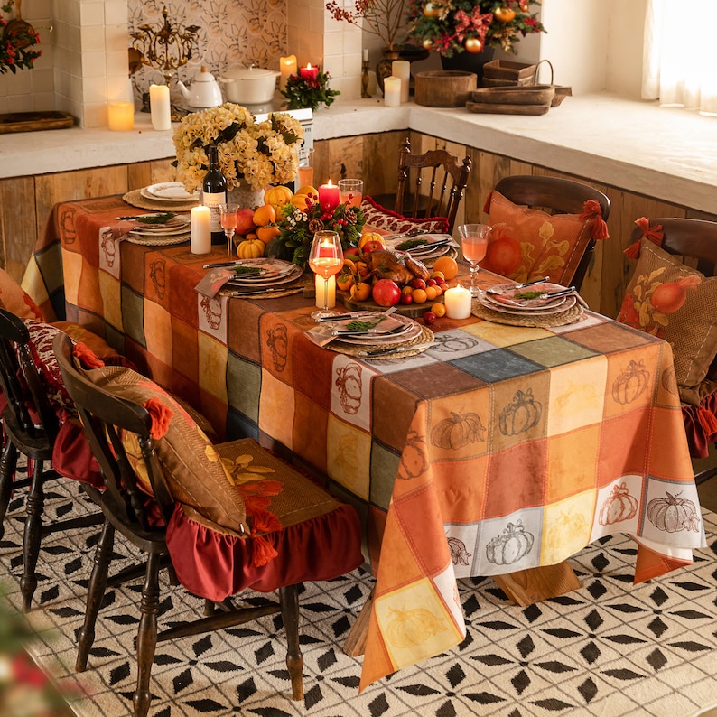 May include: A festive dining table set for a fall celebration. The table is covered with a patchwork-like tablecloth featuring pumpkin designs. Matching chair cushions and pillows add to the autumnal theme. Candles, flowers, and fruit complete the warm, inviting scene.