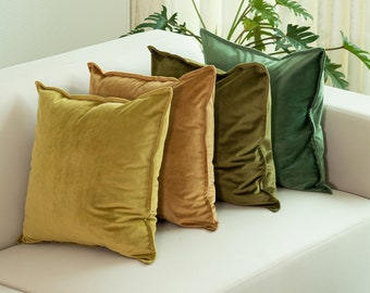May include: Four square velvet throw pillows in shades of gold, brown, and green are arranged on a white sofa. The pillows have a soft, textured appearance and are positioned against the back of the sofa, creating a layered effect.