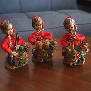 Op de afbeelding: Drie vintage keramische beeldjes van muzikanten. Elke figuur draagt een rode outfit en een bijpassende hoed. Ze spelen respectievelijk fluit, drums en een snaarinstrument. Elke figuur is omgeven door een krans van groene bladeren en rode bloemen.
