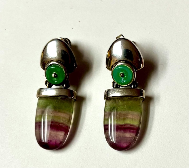 Pode incluir: Um par de brincos de prata com um detalhe circular verde e uma pedra oblonga colorida. A pedra apresenta faixas de roxo, verde e amarelo. Os brincos t&ecirc;m um acabamento met&aacute;lico polido.