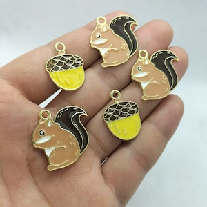 May include: Four gold-colored metal charms, two are acorn shaped with brown tops and yellow bottoms, and two are squirrel shaped with brown tails and peach-colored bodies.