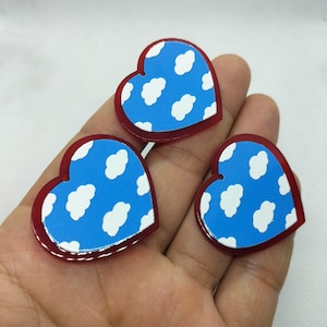 May include: Three heart-shaped charms. Each charm has a blue background with white cloud patterns, framed by a red border. The charms are held in a hand, showcasing their size and design. These charms are suitable for jewelry making or craft projects.