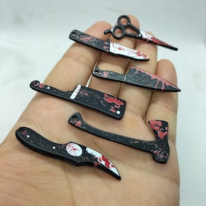 May include: A set of seven black metal keychains shaped like knives and an axe. Each keychain has a red and white paint design that resembles blood splatters.