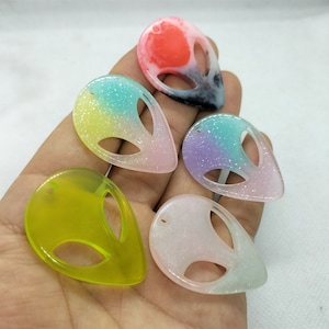 Könnte beinhalten: Fünf Alien-Kopf-förmige Harz-Anhänger mit Glitzer, der in das Harz eingebettet ist. Die Anhänger sind in verschiedenen Farben, darunter Pink, Grün, Blau, Lila und Weiß.