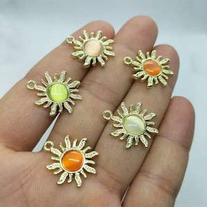 Könnte beinhalten: Fünf goldene Sonnenanhänger mit schillernden Glaszentren. Die Anhänger haben verschiedene Farben, darunter Grün, Orange und Weiß.