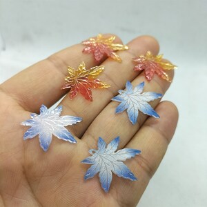 May include: Assortment of colorful maple leaf charms. The charms are made of resin and have a glossy finish. The leaves are in shades of red, orange, yellow, and blue.