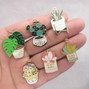 Pode incluir: Uma coleção de pingentes dourados com tema de plantas. Os pingentes apresentam várias plantas em vasos, incluindo uma folha de monstera, suculentas e cactos. Alguns pingentes têm o texto "Crazy Plant Lady", "Plant Mama" e "Plant Daddy".