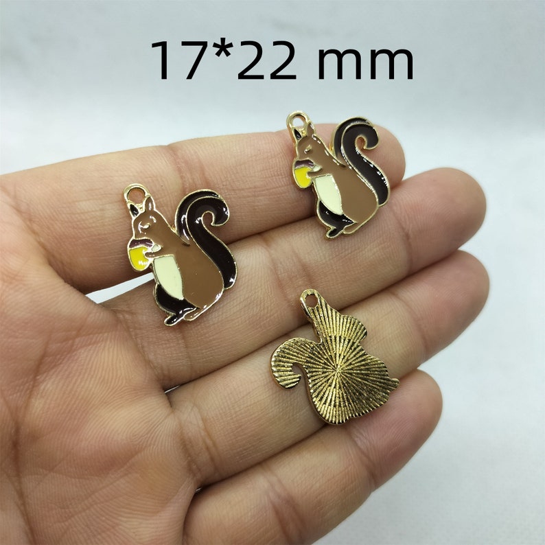 May include: Three gold-colored metal charms, each measuring 17 by 22 mm. Two charms feature a cartoon squirrel holding a nut, one with brown fur and the other with gold-colored fur. The third charm is a solid gold-colored squirrel.