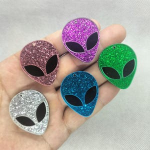 Könnte beinhalten: Eine Sammlung glitzernder Alien-Kopf-Anhänger in Rosa, Lila, Grün, Blau und Silber. Jeder Anhänger hat ein schwarzes ovales Augendesign und ein kleines Loch zum Aufhängen. Diese Anhänger eignen sich ideal für die Schmuckherstellung oder zum Basteln.