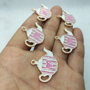 Pode incluir: Cinco pingentes de chaleira dourados com esmalte rosa e branco. Cada pingente apresenta um coração rosa e listras brancas.