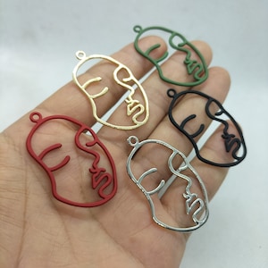 May include: Five metal face charms in different colours: gold, red, silver, black, and green. Each charm is shaped like a face with a line drawing of a face.