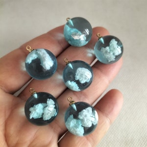 May include: Six round glass charms with gold-coloured metal loops. Each charm is filled with a light blue liquid and contains a white cloud-like shape.
