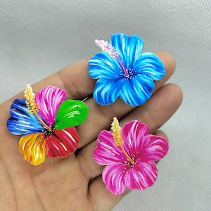 May include: Three colorful hibiscus flower charms with painted details. The flowers are blue, red, yellow, green, and pink.