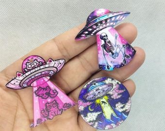 May include: Three colorful wooden UFO pins with different designs. One features a skeleton in a galaxy, another has a cat in a pink UFO, and the last one has a UFO flying over a mountain range.