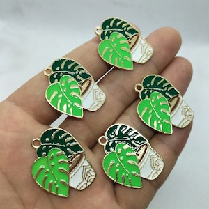 May include: Five gold-tone metal charms featuring a green and white enamel design of a potted plant with monstera leaves.