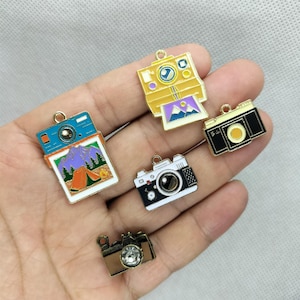 Peut inclure: Une collection de breloques en forme d'appareils photo colorés. Les breloques comprennent un appareil photo instantané bleu et blanc avec une scène de montagne, un appareil photo instantané jaune, un appareil photo noir et or, un appareil photo noir et blanc et un appareil photo marron avec un cristal.