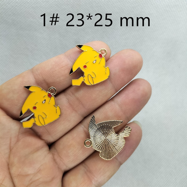 May include: Three gold-colored metal charms. Two charms feature a yellow cartoon character with red cheeks and black eyes. The third charm is a gold-colored, textured, round shape. The charms are 23 mm by 25 mm.