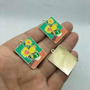 May include: Two gold-toned metal charms with a green background and a painting of sunflowers in a vase. One charm is blank with a sunburst design.