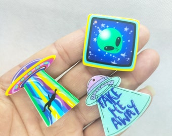 May include: Three colorful alien-themed pins. One pin features a green alien face on a blue background. Another shows a UFO with a beam and a silhouetted figure. The third pin has a UFO with the text "TAKE ME AWAY".