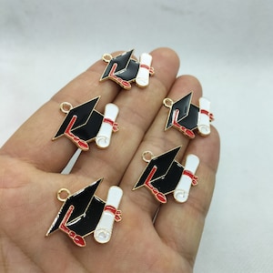 Puede incluir: Cinco dijes de gorro de graduación de color dorado con un rollo blanco y detalles rojos. Los dijes están en una mano.