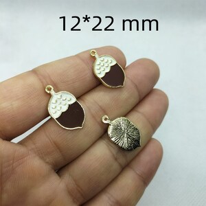 May include: Two gold-tone acorn charms with white and brown enamel. The charms are 12 mm by 22 mm.