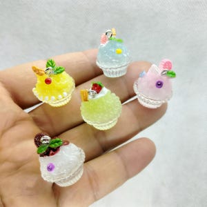 Könnte beinhalten: Fünf Miniatur-Dessert-Anhänger in verschiedenen Farben. Jeder Anhänger hat eine andere Farbe, darunter Gelb, Grün, Blau, Rosa und Weiß. Die Anhänger sind mit kleinen Details wie Früchten und Blättern verziert und stehen auf einer weißen Basis.
