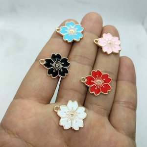 May include: Five gold-tone metal flower charms with enamel paint in different colors: black, white, red, pink, and blue. Each charm has a loop for attaching to jewelry or other crafts.