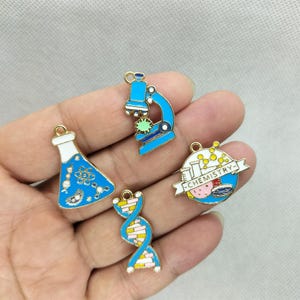 Könnte beinhalten: Vier wissenschaftliche Anhänger in Blau, Weiß und Gold. Enthält ein Mikroskop, einen Kolben, eine DNA-Strähne und einen runden Anhänger mit dem Wort "CHEMISTRY". Jeder Anhänger hat eine goldfarbene Schlaufe zum Befestigen.