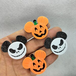 May include: Four Halloween-themed ornaments, two are orange pumpkins with black eyes and a black mouth, and two are black and white with a white skull and black eyes and mouth. The ornaments are shaped like Mickey Mouse ears.