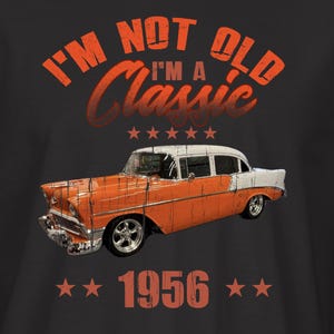 May include: Black t-shirt featuring a classic orange and white car graphic with the text "I'M NOT OLD I'M A Classic" above the car and "1956" below. The shirt has a small emblem on the left sleeve.