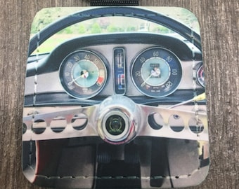 May include: A square leather keychain with a photo of a vintage car's dashboard. The photo shows the steering wheel, speedometer, and other gauges. The keychain has a black strap and a silver key ring.