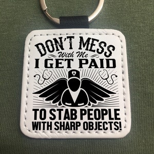 Könnte beinhalten: Weißer Leder-Schlüsselanhänger mit schwarzem Text, der "Don't Mess With Me I Get Paid To Stab People With Sharp Objects!" lautet. Der Schlüsselanhänger zeigt eine schwarze Silhouette einer Krankenschwester mit Flügeln und einem Stethoskop.