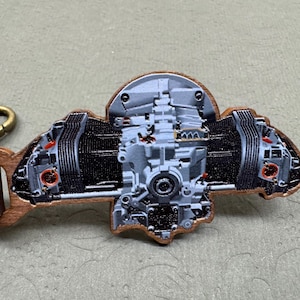 May include: A wooden keychain featuring a detailed, gray and black engine design. The engine has red accents and is attached to a brown leather strap with a gold-colored clasp. The keychain is set against a neutral background.