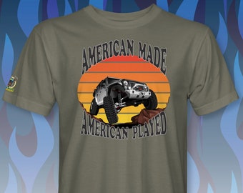 May include: A green t-shirt with a graphic of a silver Jeep driving over rocks. The graphic has a sunset background and the text "American Made American Played".