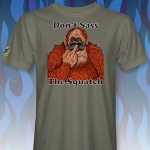 Peut inclure: Un t-shirt vert avec une image de dessin anim&eacute; d'un sasquatch portant des lunettes de soleil et faisant un doigt d'honneur. Le texte "Don't Sass The Squatch" est imprim&eacute; sur le t-shirt.