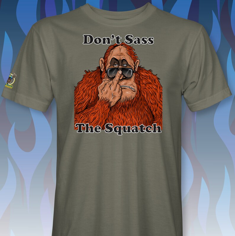 Peut inclure: Un t-shirt vert avec une image de dessin anim&eacute; d'un sasquatch portant des lunettes de soleil et faisant un doigt d'honneur. Le texte "Don't Sass The Squatch" est imprim&eacute; sur le t-shirt.