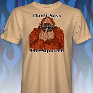 Peut inclure: Un t-shirt beige avec une image de dessin anim&eacute; d'un sasquatch portant des lunettes de soleil et faisant un doigt d'honneur. Le texte "Don't Sass The Squatch" est imprim&eacute; sur le t-shirt.