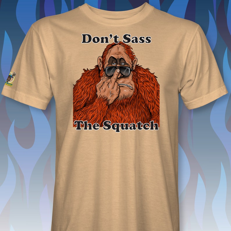 Peut inclure: Un t-shirt beige avec une image de dessin anim&eacute; d'un sasquatch portant des lunettes de soleil et faisant un doigt d'honneur. Le texte "Don't Sass The Squatch" est imprim&eacute; sur le t-shirt.