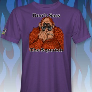 Peut inclure: T-shirt violet avec une image de dessin anim&eacute; d'un sasquatch portant des lunettes de soleil et faisant un doigt d'honneur. Le texte "Don't Sass The Squatch" est imprim&eacute; sur le t-shirt.