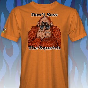 Peut inclure: T-shirt orange avec une illustration de dessin anim&eacute; d'un sasquatch portant des lunettes de soleil et faisant un doigt d'honneur. Le texte "Don't Sass The Squatch" est imprim&eacute; sur le t-shirt.