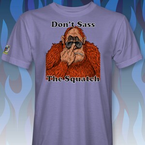 Peut inclure: T-shirt violet avec une image de dessin anim&eacute; d'un sasquatch portant des lunettes de soleil et faisant un doigt d'honneur. Le texte "Don't Sass The Squatch" est imprim&eacute; sur le t-shirt.