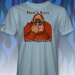 Peut inclure: Un t-shirt bleu clair avec une illustration de dessin anim&eacute; d'un sasquatch portant des lunettes de soleil et faisant un doigt d'honneur. Le texte "Don't Sass The Squatch" est imprim&eacute; sur le t-shirt.
