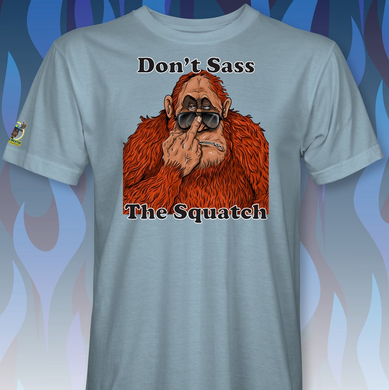 Peut inclure: Un t-shirt bleu clair avec une illustration de dessin anim&eacute; d'un sasquatch portant des lunettes de soleil et faisant un doigt d'honneur. Le texte "Don't Sass The Squatch" est imprim&eacute; sur le t-shirt.