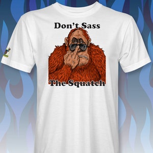 Peut inclure: T-shirt blanc avec une image de dessin anim&eacute; d'un sasquatch portant des lunettes de soleil et faisant un doigt d'honneur. Le texte "Don't Sass The Squatch" est imprim&eacute; sous l'image.